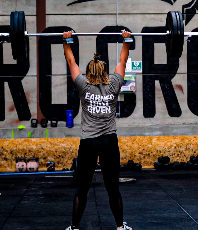 Mujer de espaldas levantando una barra con pesas sobre su cabeza en un gimnasio.